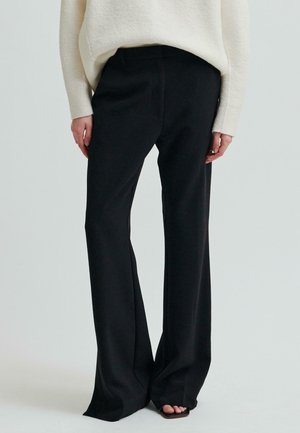 Žena oblečená v černých širokých kalhotách s vysokým pasem a bílém svetru s dlouhými rukávy, stojící bosky na jednobarevném pozadí.