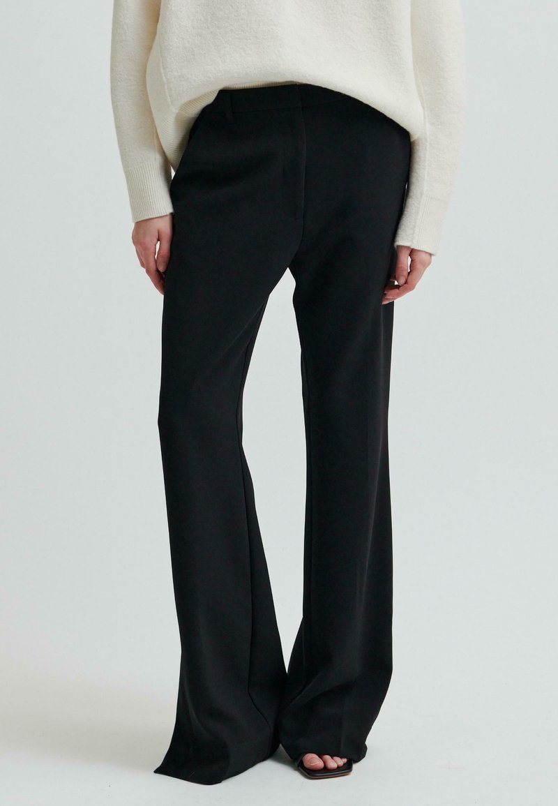 Kobieta w czarnych szerokich spodniach z wysokim stanem i białym swetrze z długim rękawem, stojąca boso na jednolitym tle.