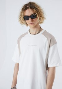 White t-shirt with beige raglan sleeves, featuring a logo in light gray. Material appears soft and lightweight. Chain necklace visible.