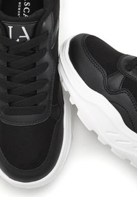 Sneakers neri realizzati con un mix di pelle sintetica e camoscio, caratterizzati da una suola bianca spessa e cuciture dettagliate sulla tomaia.