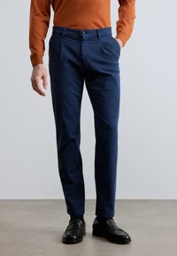 Marinblå plisserade byxor i bomull med knappstängning, sidfickor och avsmalnande ben, kompletterade med svarta loafers.