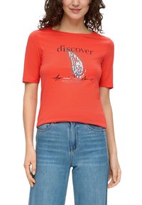 Czerwona koszulka z krótkim rękawem z dekoltem w łódkę, z graficznym wzorem tekstu i kolorowym motywem. Noszona z jasnoniebieskimi dżinsami.