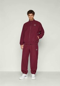 Bordo treniņtērps ar ar rāvējslēdzēju un elastīgiem biksēm, kurām kreisajā krūtī ir logo, kā arī tāds pats logo uz biksēm. Baltas kedas.