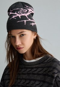 Beanie grigio lavorato a maglia con un design di rosa rosa e spine, indossato con un maglione scuro e strutturato con motivo a treccia e colletto bianco.
