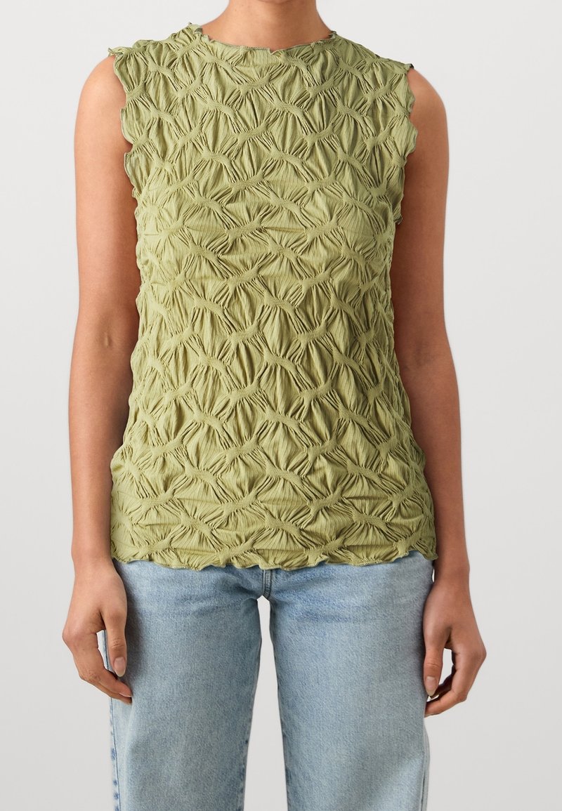 Haut sans manches de couleur vert olive texturé avec un motif en losanges froncés porté avec un jean bleu clair par une personne aux bras relaxés.