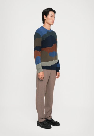 Pulover tricotat cu modele abstracte în albastru, verde, brun și bleumarin; textură poziționată. Asortat cu pantaloni bej și pantofi negri.