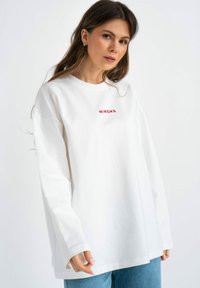 Witte long-sleeve shirt met rode geborduurde tekst "MIRONS." Gemaakt van katoen met een ontspannen pasvorm en ronde hals. Gemaakt in combinatie met lichtblauwe jeans.