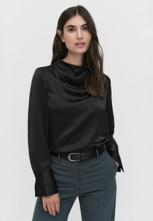Zwarte satijnen blouse met een halslijn met plooien en lange mouwen met brede manchetten. Gepaard met grijze, op maat gemaakte broeken en een zwarte riem.