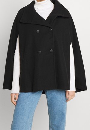 Cappotto nero doppiopetto con grandi bottoni, colletto ampio e vestibilità ampia. Indossato sopra un dolcevita bianco a coste e jeans blu.
