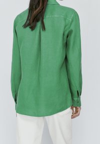 Person wearing a long-sleeve green shirt and white pants, shown from the back against a plain light background.