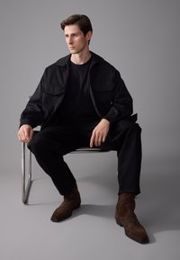 Chaqueta negra con dos bolsillos en el pecho sobre una camiseta negra, combinada con pantalones negros y botas de ante marrón. Modelo sentado en una silla de cromo.