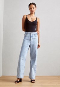 Svart bodysuit med tunna axelband i kombination med högmidjade ljusblå raka jeans och svarta klacksandaler. Neutral bakgrund.