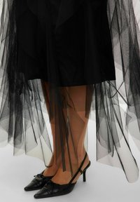 Schwarzer, transparenter Tüllrock, der über einem einfarbigen schwarzen Unterrock geschichtet ist, kombiniert mit schwarzen Slingback-Absätzen mit spitzem Zehen und Schleifendetail.