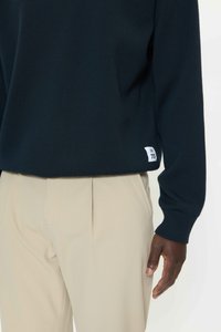 Mornarsko modra pulover jopica z rebrastimi manšetami in spodnjim robom, kombinirana z bež hlačami. Na spodnjem levem delu je majhna etiketa s številko 73.