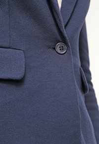 Blazer Navy en tissu texturé, avec une fermeture à un seul bouton et une poche à rabat inclinée sur le côté gauche.