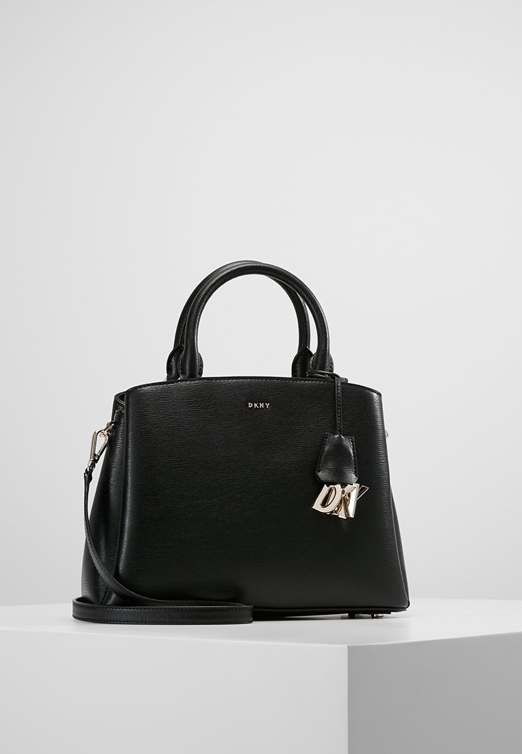 Sac à main DKNY en cuir noir avec doubles poignées, bandoulière détachable et breloque logo DKNY dorée sur un socle blanc.