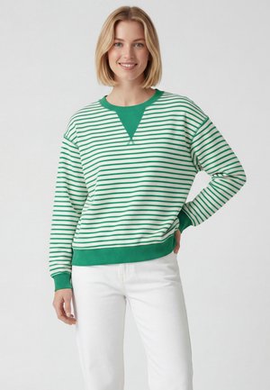 Giovane donna con capelli biondi corti che indossa un maglione a righe verdi e bianche e pantaloni bianchi, sorridente con una mano sui fianchi.