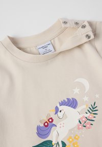 Crème peuter T-shirt met drukknoopjes op de schouder, versierd met een eenhoorn met paarse manen, vlinders, bloemen, een maan en sterren.