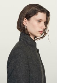 Czarny wełniany płaszcz z wysokim kołnierzem, zapięciem na jeden guzik oraz gładką fakturą. Model ma delikatne fale we włosach.