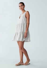 Biała sukienka na falbanach z cienkimi niebieskimi paskami, dekoltem w serek, bez rękawów i rozszerzanym dołem. Modelka ma na sobie płaskie sandały.