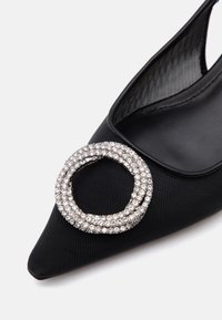 Chaussure plate noire à bout pointu avec une grande boucle circulaire en strass sur la partie supérieure avant, sur fond blanc.