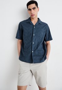 Camisa de manga corta hecha de tela de patrón azul oscuro, con cuello de campamento, cierre de botones en el frente y un bolsillo en el pecho. Combinada con pantalones cortos beige.
