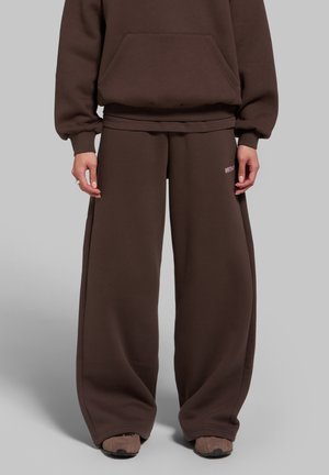 Personne portant un pantalon de jogging marron à jambes larges et un sweat à capuche marron assorti, debout sur un fond gris uni.