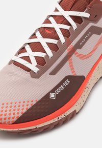 Gros plan sur une chaussure Nike Trail beige et rouille avec des lacets blancs, une étiquette Gore-Tex mise en avant et une semelle avec accents orange et une semelle intermédiaire tachetée.