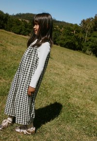 Jeune fille aux longs cheveux noirs portant une chemise blanche à manches longues et une robe à carreaux noir et blanc, debout dans un champ herbeux avec des arbres en arrière-plan.