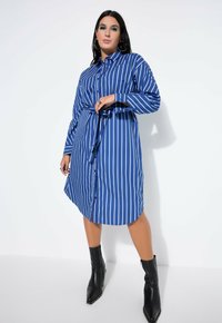 Robe chemise rayée bleue et blanche avec manches longues, boutons sur le devant, ceinture à la taille, et bottines noires unies. Tissu lisse avec une coupe décontractée.