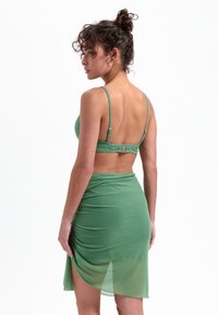 Zielony dwuczęściowy zestaw składający się z dopasowanego biustonosza na ramiączkach z regulacją oraz pasującą, przezroczystą spódnicą z bocznym rozcięciem.