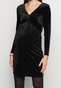 Gravid kvinna som bär en svart sammetsklänning med lång ärm och åtsittande passform, med en tvinnad knutdetalj vid bröstet.