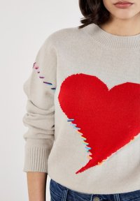 Beżowy sweter w dzianinę z dużym czerwonym sercem na przodzie, ozdobiony wielokolorowymi przeszyciami wzdłuż lewego rękawa i u dołu.