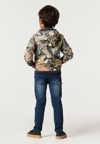 Kamo-mönstrad huva-jacka i gröna, bruna och blå bladverk färger. Kombinerad med blå denimjeans och svarta skor med beige sulor.