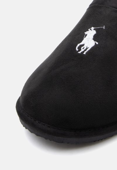 Slipper em camurça preta com logotipo de jogador de polo branco bordado na parte superior da frente e borda da sola de borracha preta visível.