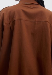 Chemise en lin marron avec une coupe décontractée, dotée d'un col classique, d'épaulettes sur les épaules et d'une discrète broderie de logo à l'arrière.