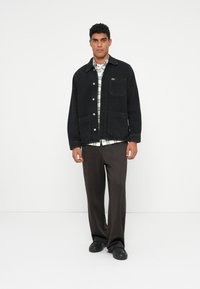 Giacca in denim nera con chiusura frontale a bottoni, due tasche grandi e un piccolo dettaglio con logo. Abbinata a una camicia a quadri e pantaloni marroni.