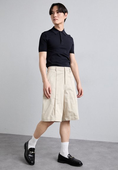 Polo shirt noir à manches courtes, shorts plissés beiges arrivant aux genoux et mocassins noirs associés à des chaussettes blanches. Texture de tissu lisse.