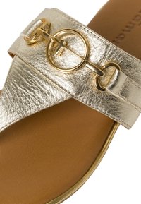 Metallisk guld slide sandal med en bred rem som har en cirkulär guldaccent. Slät textur med en tan-färgad fotbädd och en lätt glans.