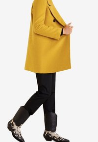 Geel wollen jas met een double-breasted ontwerp, gecombineerd met zwarte broek en zwarte cowboylaarzen met witte sierlijke details.