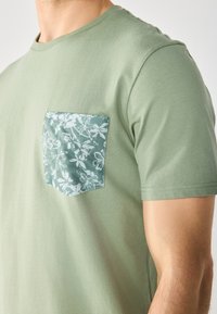 Green cotton t-shirt with a crew neck, featuring a floral-patterned pocket in a darker green. Smooth texture, short sleeves.