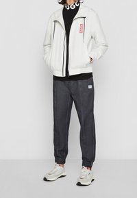 Bílá lehká bunda s kapucí a černým zipem, kombinovaná se šedými kalhotami ve stylu jogger. Obsahuje červené logo, elastické manžety a bílé tenisky.