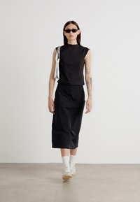 Svart ermeløs topp og lang skjørt, begge laget av glatt stoff. Modellen har på seg svarte solbriller, hvite sokker og lysegrå sneakers.