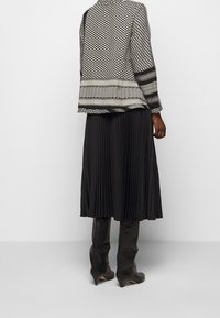 Schwarzer gefalteter Rock kombiniert mit einer geometrisch gemusterten Jacke in Schwarz- und Weißtönen, langen Ärmeln und ausgeprägten Stoffstrukturen.