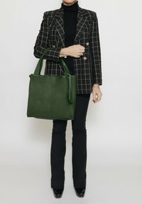 Borsa tote in pelle verde con doppio manico, forma rettangolare, texture liscia e un pendente con nappina staccabile. Blazer a quadri nero e oro.