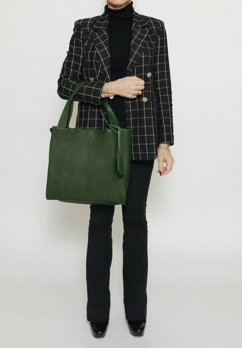 Borsa tote in pelle verde con doppio manico, forma rettangolare, texture liscia e un pendente con nappina staccabile. Blazer a quadri nero e oro.
