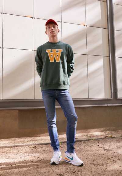 Grüner Kapuzenpullover mit gelbem "W"-Logo, blaue Jeans und mehrfarbige Nike-Sneaker. Der Hintergrund besteht aus hellen Paneelen und einem strukturierten Boden.