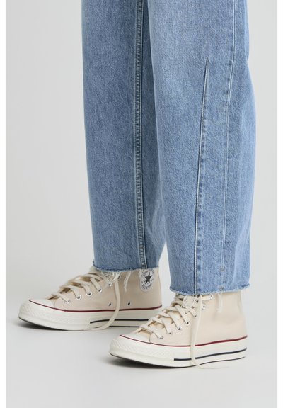Zapatillas altas de lona beige con suelas de goma blancas y detalles en rojo, combinadas con unos vaqueros de denim azul claro con dobladillos deshilachados.