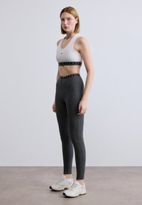 Λευκό αθλητικό σουτιέν με μαύρη ταινία, γκρι κολάν και μπεζ αθλητικά παπούτσια με μαύρη σόλα, παρουσιάζοντας εφαρμοστή σιλουέτα και λεία υφή.