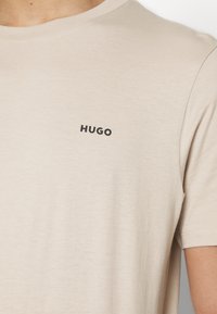 T-shirt in cotone beige con scollatura rotonda e maniche corte. Presenta un logo nero "HUGO" sul lato sinistro del petto. Texture morbida.